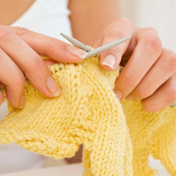 A close up image of a woman's hands knitting a lightweight garment for summer out of a pale yellow wool yarn.
